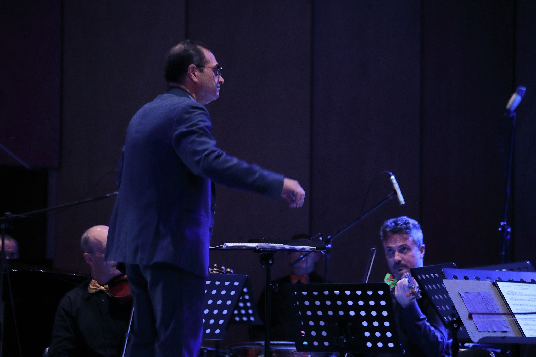 Il M° Carmelo Chillemi dirige l'orchestra — ripresa di profilo, luce blu scenografica