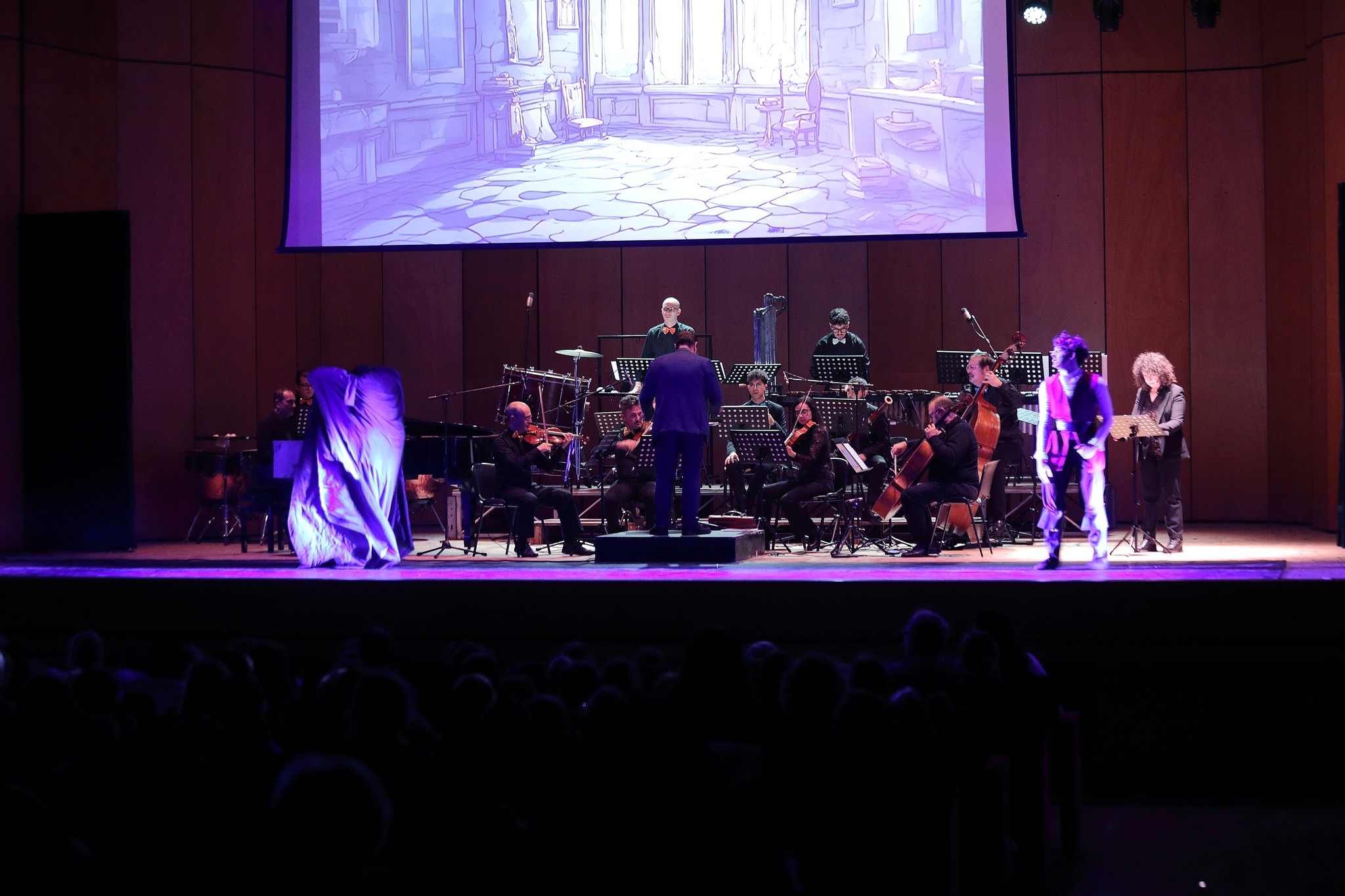 Orchestra e personaggi in scena — stanza medievale proiettata, vista dalla platea 2024