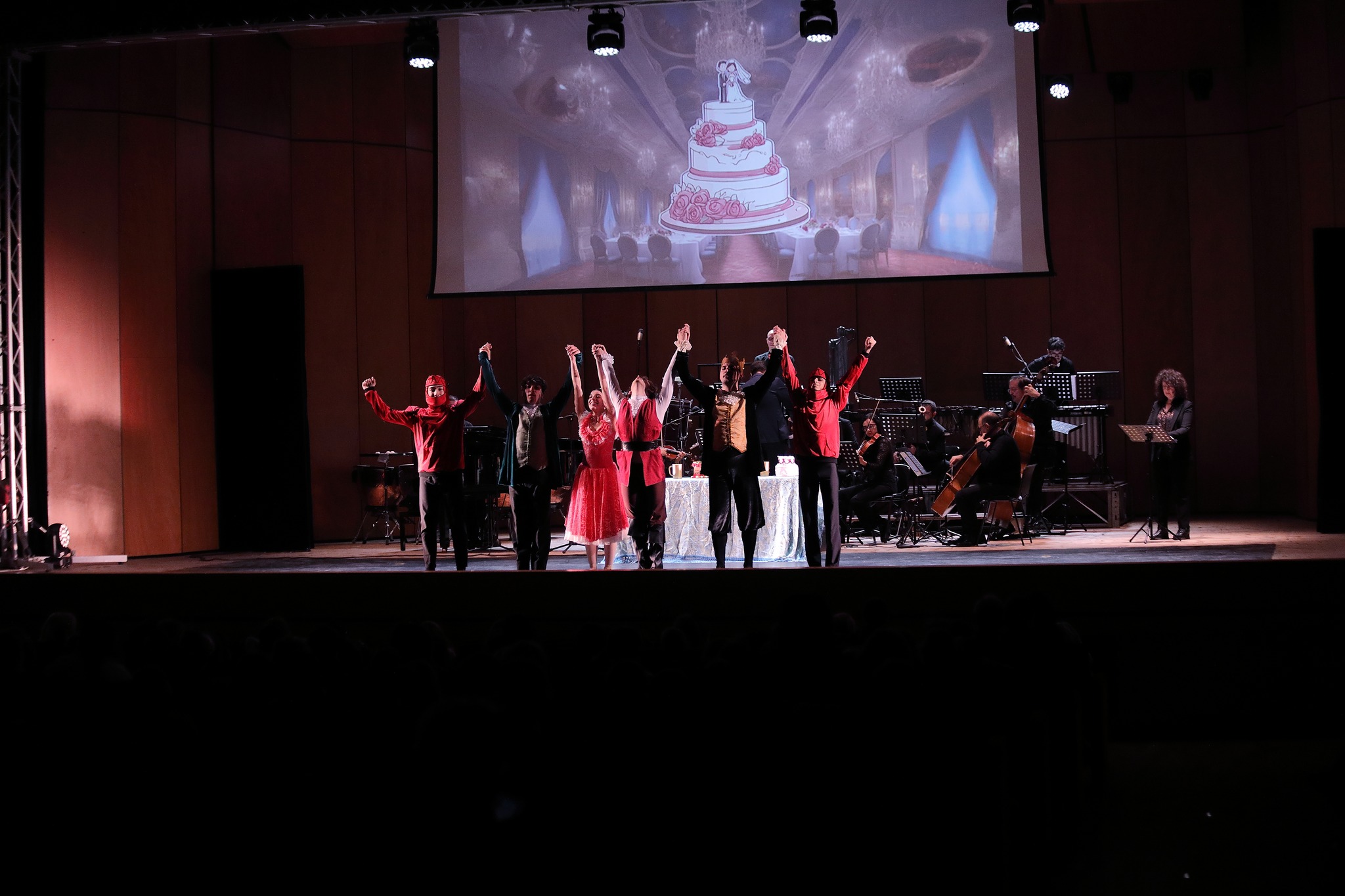 Il cast unisce le mani alzate — finale con la torta nuziale sullo schermo, orchestra 2024